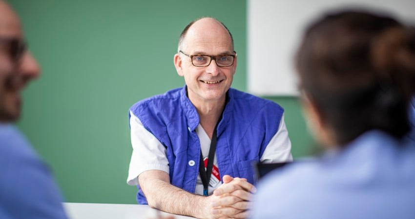 Frieder Braunschweig sitter i blå/vita sjukhuskläder och svarta glasögon leendes. Han sitter i ett grönt rum.