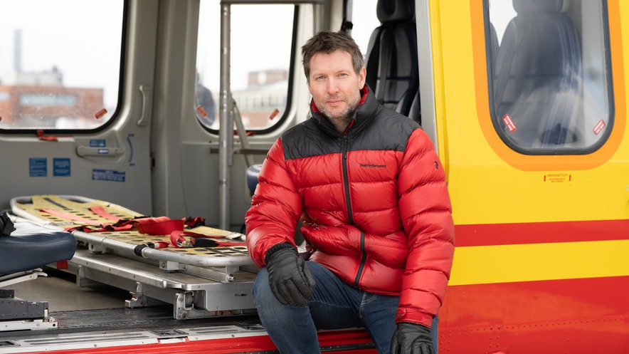 Lars Falck i rödsvarta kläder sitter i helikoptern med en grågul bår.