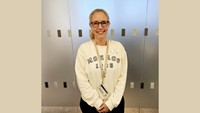 Anna har en beige sweatshirt på sig och står framför en glasvägg på sjukhuset.