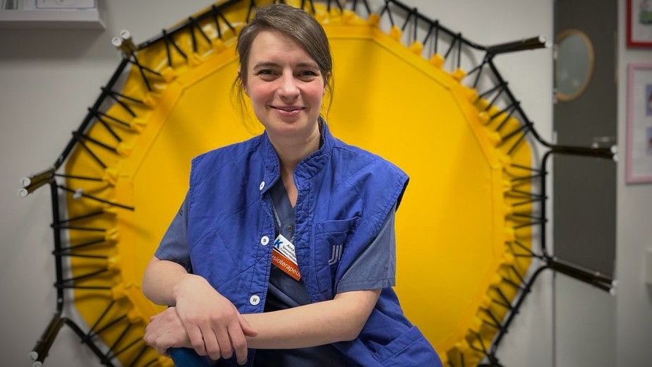 Sjukgymnasten Anna Hedborg sitter i blå arbetskläder framför en gul studsmatta.