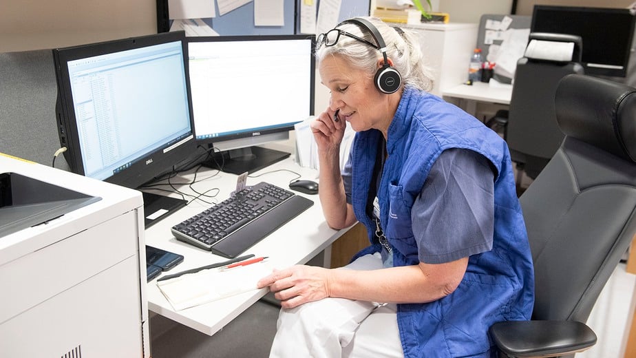 Sjuksköterska i blå väst och vita byxor pratar i telefon vid sitt skrivbord.