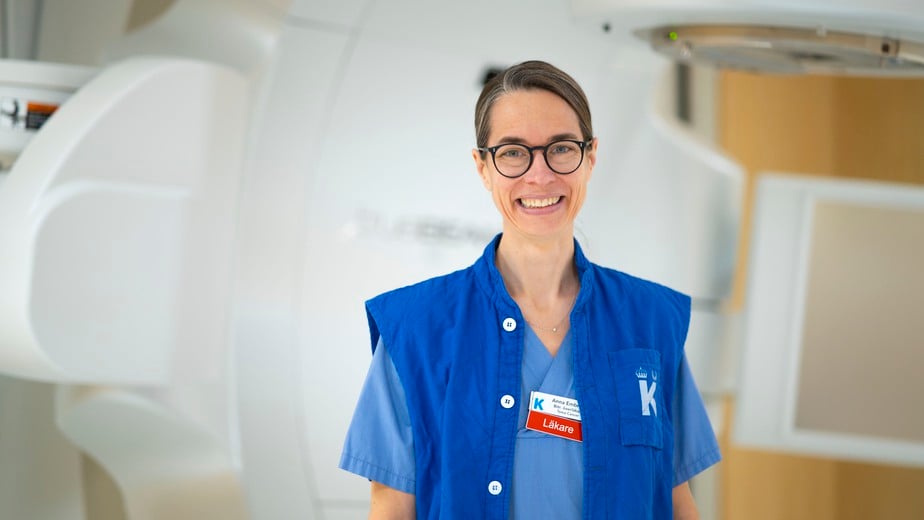 Anna Embring i sjukvårdskläder framför en strålningsapparat, hon ler och tittar in i kameran