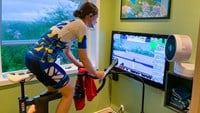 Anna Embring på motionscykel i hemmet framför en stor tv som visar en animerad värld