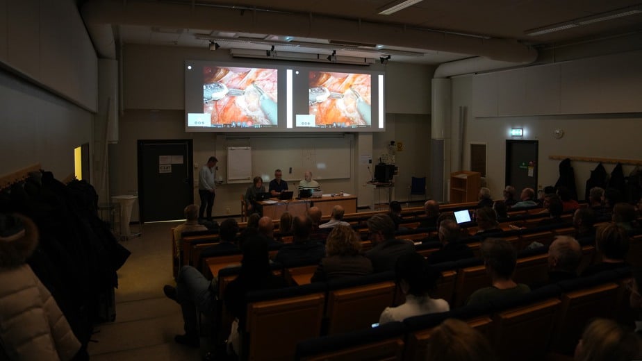 80 personer sitter i rader i en aula och tittar på två stora bildskärmar.