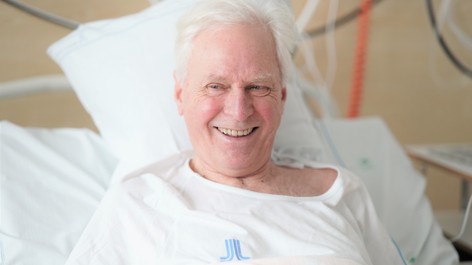 Patient Björn Persson ler i sängen med  vita sjukhuskläder