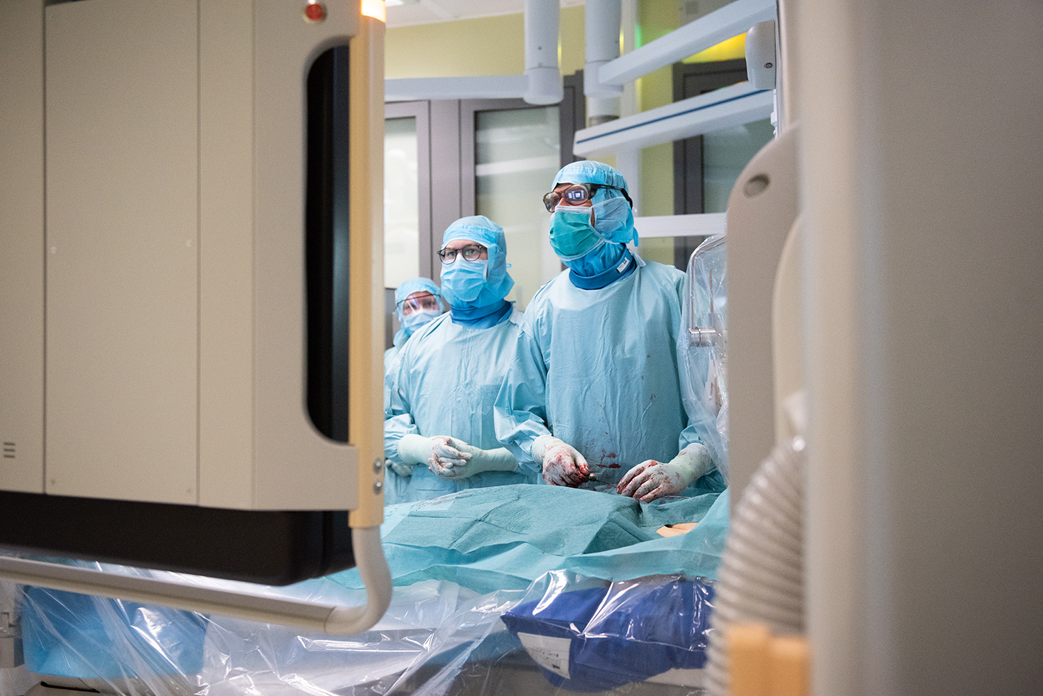 Bakom en stor skärm står två operationsbeklädda kärlkirurger framför ett operationsbord