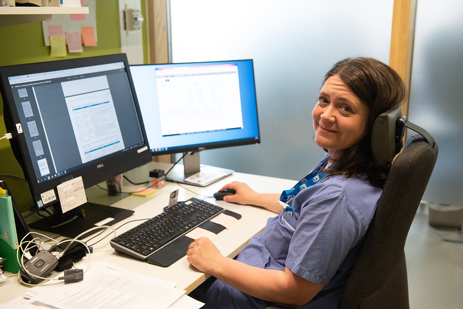 Forskningssjuksköterska sitter vid skrivbordet med datorskärmar och ler mot kameran