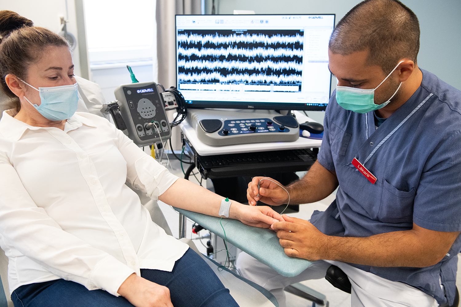 Läkare placerar en nål i sidan av handen på en patient, en skärm i bakgrunden visar dom elektriska signalerna