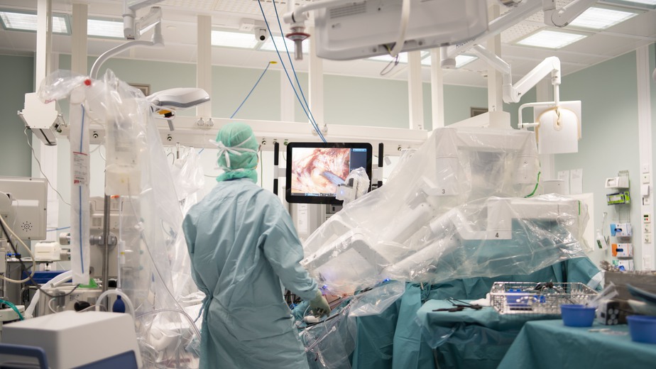 En kirurg står intill en stor flerarmad robot inne i ett operationsrum.