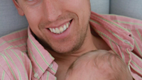 En man tittar leendes in i kameran, ett nyfött barn sover i hans famn
