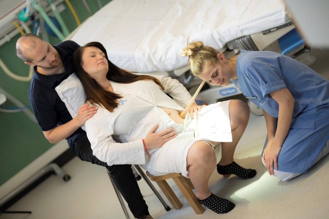 kvinna sitter på förlossningspall med partner bakom och barnmorskan lyssnar med tratt