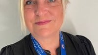 Anna har blont hår uppsatt i en tofs och en svart blus på sig.