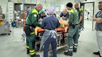 Cechael står i blå sjukhuskläder och ett plastförkläde och assisterar ett akutteam.