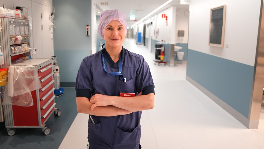 Cechael Sebghati Sparrfelt är klädd i blå sjukhuskläder och en rosa operationsmössa. Hon står i en lång ljus sjukhuskorridor.
