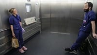 Cechael åker hiss med kollegan Johan. Båda har blå arbetskläder och röda läkarskyltar. De skrattar.