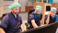 Cechael och kollegorna Gustaf och Johan sitter och samtalar i blå arbetskläder och gröna operationsmössor vid en stor dator på kontoret.