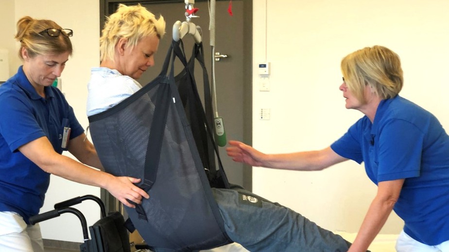 Två arbetsterapeuter visar hur man kan lyfta upp en patient i en sittlift.