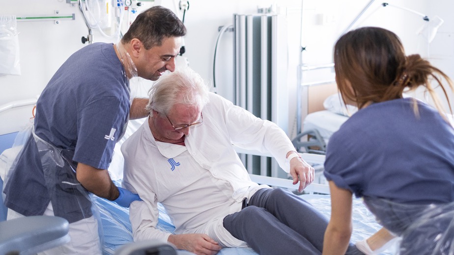 Två sjuksköterskor hjälper en äldre man upp ur sängen.