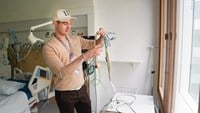 En ung man står vid fönstret i ett patientrum och visar upp en knippe kablar för patientövervakning.