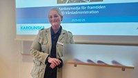 Kvinna i blå jeans och beige jacka står framför en whiteboard.