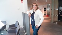 Kvinna i blå jeans och vit kofta står med armen lutad mot en grå skärmvägg.