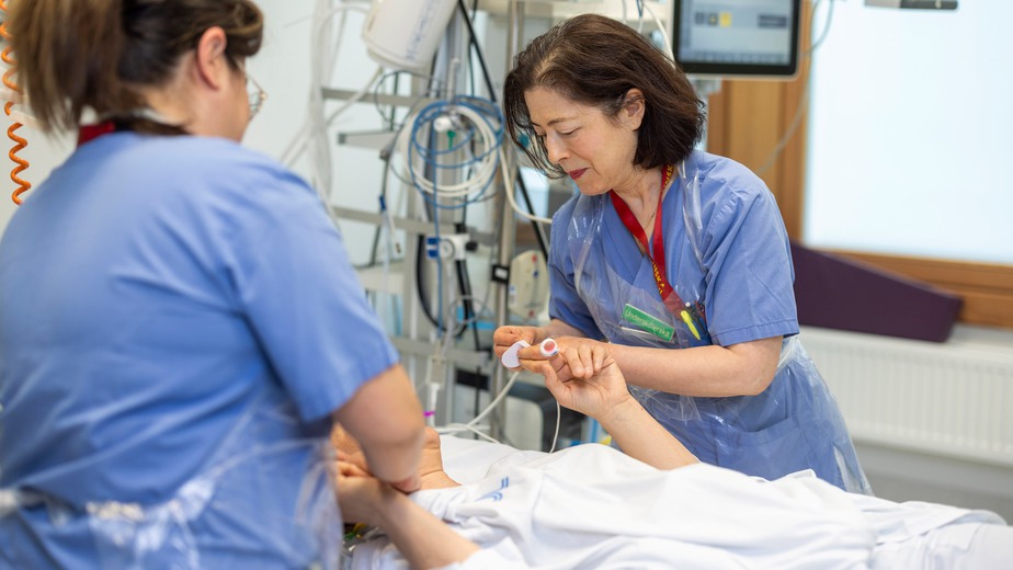Sjuksköterska och undersköterska tar hand om en patient som ligger i en sjukhussäng.
