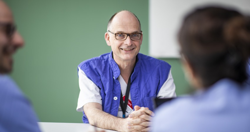Frieder Braunschweig sitter i blå/vita sjukhuskläder och svarta glasögon leendes. Han sitter i ett grönt rum.