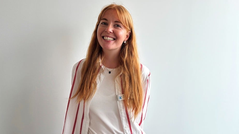 Nora Mårtensson har rött långt hår, vitrödrandig skjorta och svarta jeans. Hon står mot en vit vägg och ler in i kameran.