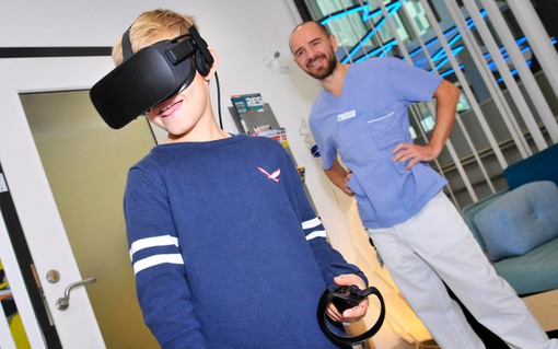 Ett barn testar virtual reality och har på sig vr-mask och håller i en handkontroll samtidigt som en vårdpersonal tittar på leende