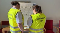 Två personer i gula västar som står framför ett bord.