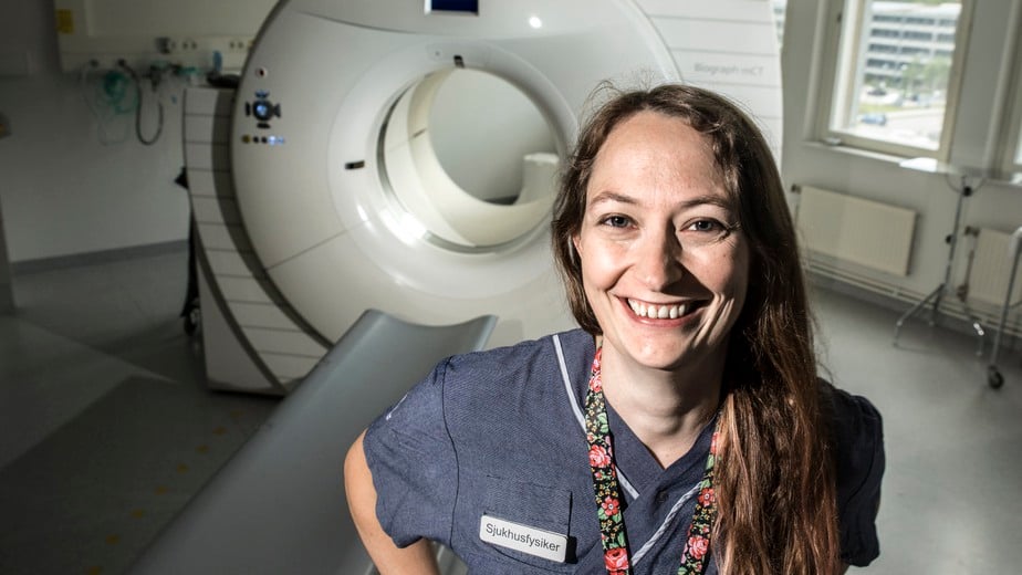 Sjukhusfysiker Maria Holstensson står framför en stor vit petkamera och ler brett