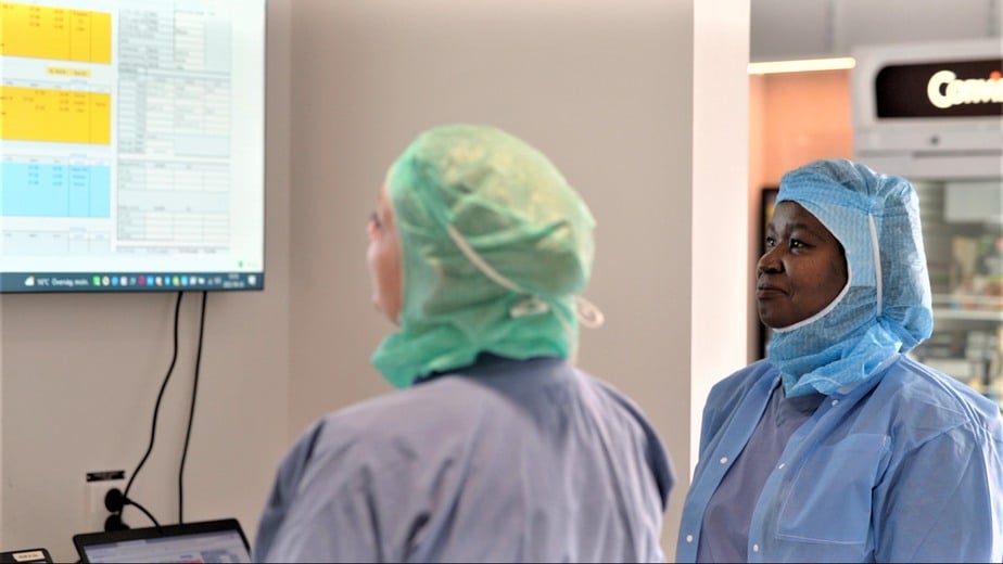 Sjuksköterskorna Saralena Johansson och Anna Mwaka står i korridoren och tittar på en bildskärm som visar lördagens operationsschema.