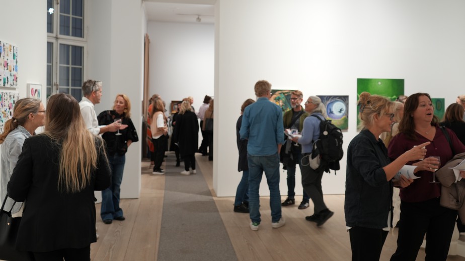 Ett galleri med två rum och med höga, vita väggar fullt med människor som minglar och tittar på tavlor som hänger uppe på väggarna