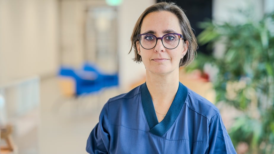 Emilia har blå sjukhusskjorta och svarta glasögon med lila detaljer. Hennes hår är klippt i en kort page.