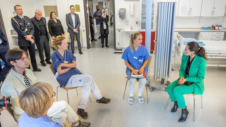 Kronprinsessan i grönsvart sitter på en stol och talar med 3 läkare i blåkläder samt en patient i vit kostym.