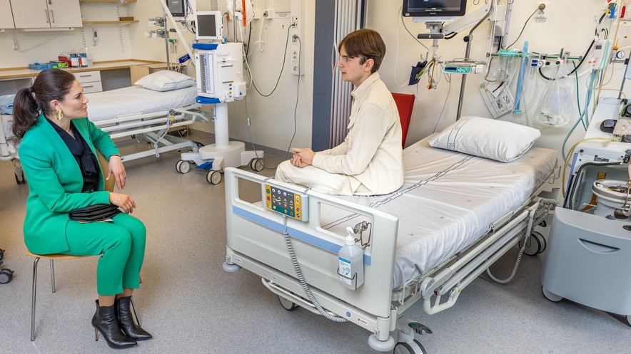 Kronprinsessan i grönsvart sitter på en stol och talar med en patient i vit kostym som sitter på en säng.