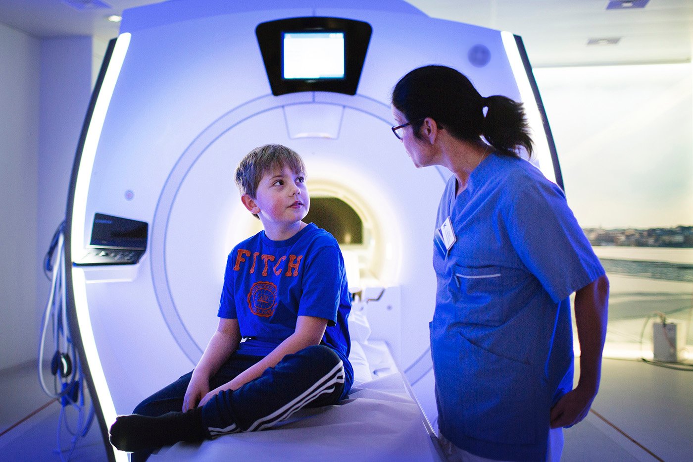bilden visar en pojke som sitter framför en magnetröntgenkamera. Bredvid pojken står en kvinna i sjukvårdsklädsel. Hon har glasögon och tittar på pojken.
