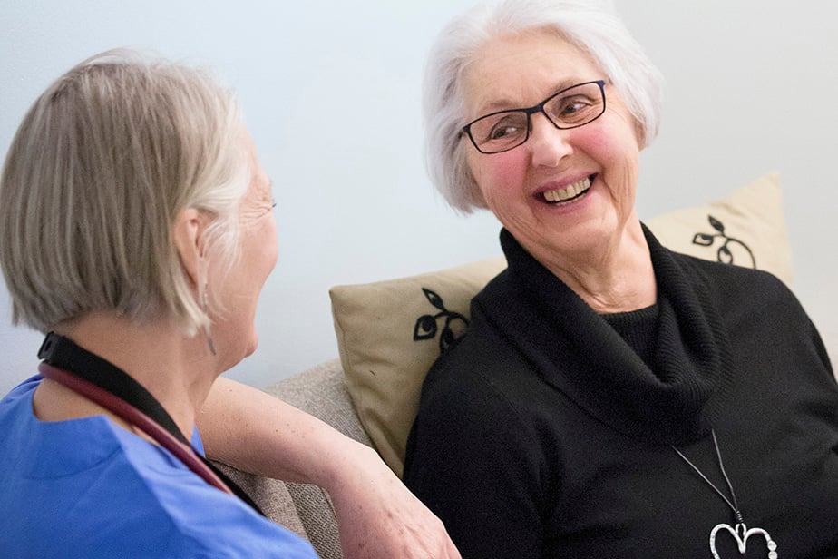 bilden visar en äldre kvinna i vitt hår som ler och tittar på en annan kvinna med band rund halsen och blå skjorta i sjukvårdsklädsel.
