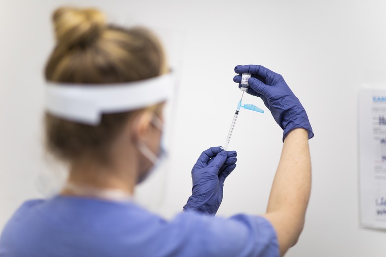 Vaccination mot covid-19. Sjuksköterska i visir och med munskydd och handskar drar upp vaccin i spruta.