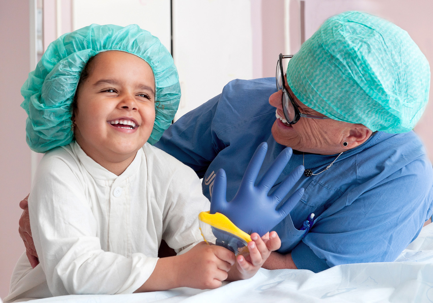 En vårdklädd kvinna lutar sig in bredvid ett skrattande barn klädd i sjukhuskläder och operationshuva.