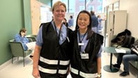 En blond kvinna i kortklippt hår står bredvid en kvinna i långt brunt hår och asiatiskt utseende. De har på sig sjukhusets svarta stabsvästar.