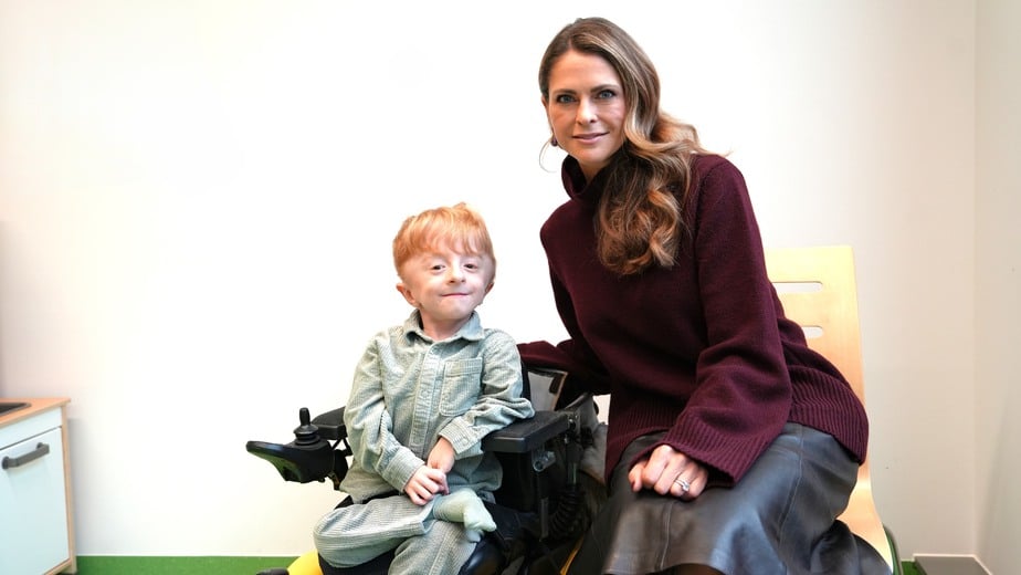 Oliver sitter i sin elrullstol intill prinsessan Madeleine. De ler in i kameran.