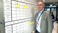 Medelålders man med mycket kort hår och rektangulärara glasögon klädd i blå jeans och grå kavaj. Han står vid en stor whiteboard med postitlappar.