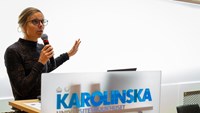 Josefin Wångdahl med en mikrofon framför ett podium med texten Karolinska Universitetssjukhuset och vit, ljus bakgrund.