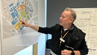 En blond man i svarta stabskläder pekar på en karta över Karolinska Universitetssjukhuset.
