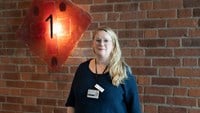 Kvinna står framför en tegelvägg och tittar in i kameran.