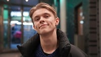 Theoz har blont vågit hår och är klädd i svart täckjacka.