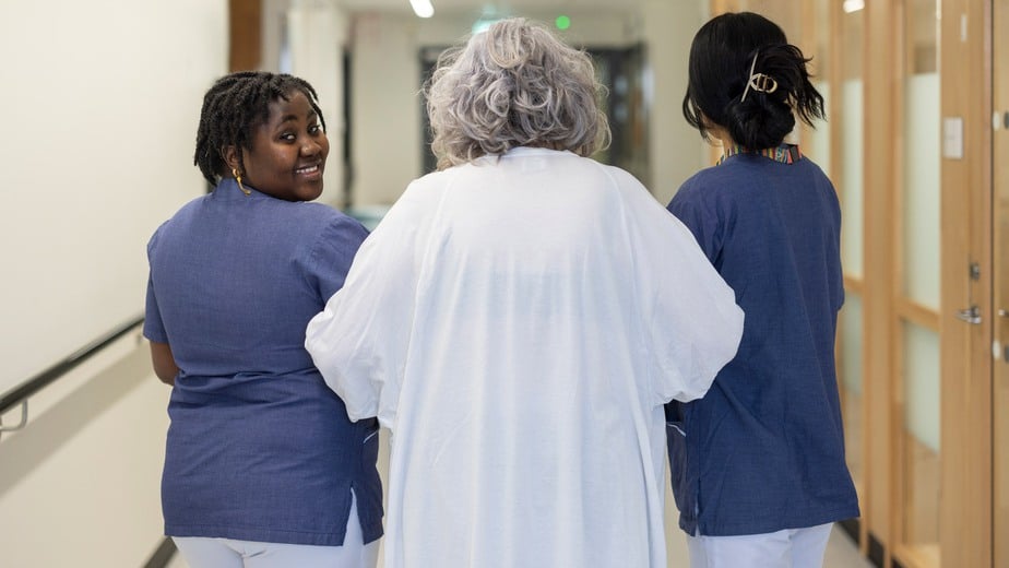 En äldre kvinna i vita sjukhuskläder får hjälp av två sköterskor att ta sig fram i en korridor.