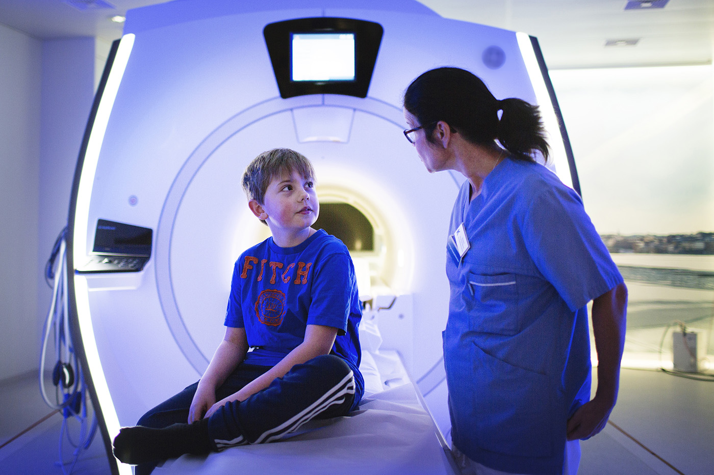 bilden visar en pojke som sitter framför en magnetröntgenkamera. Bredvid pojken står en kvinna i sjukvårdsklädsel. Hon har glasögon och tittar på pojken.